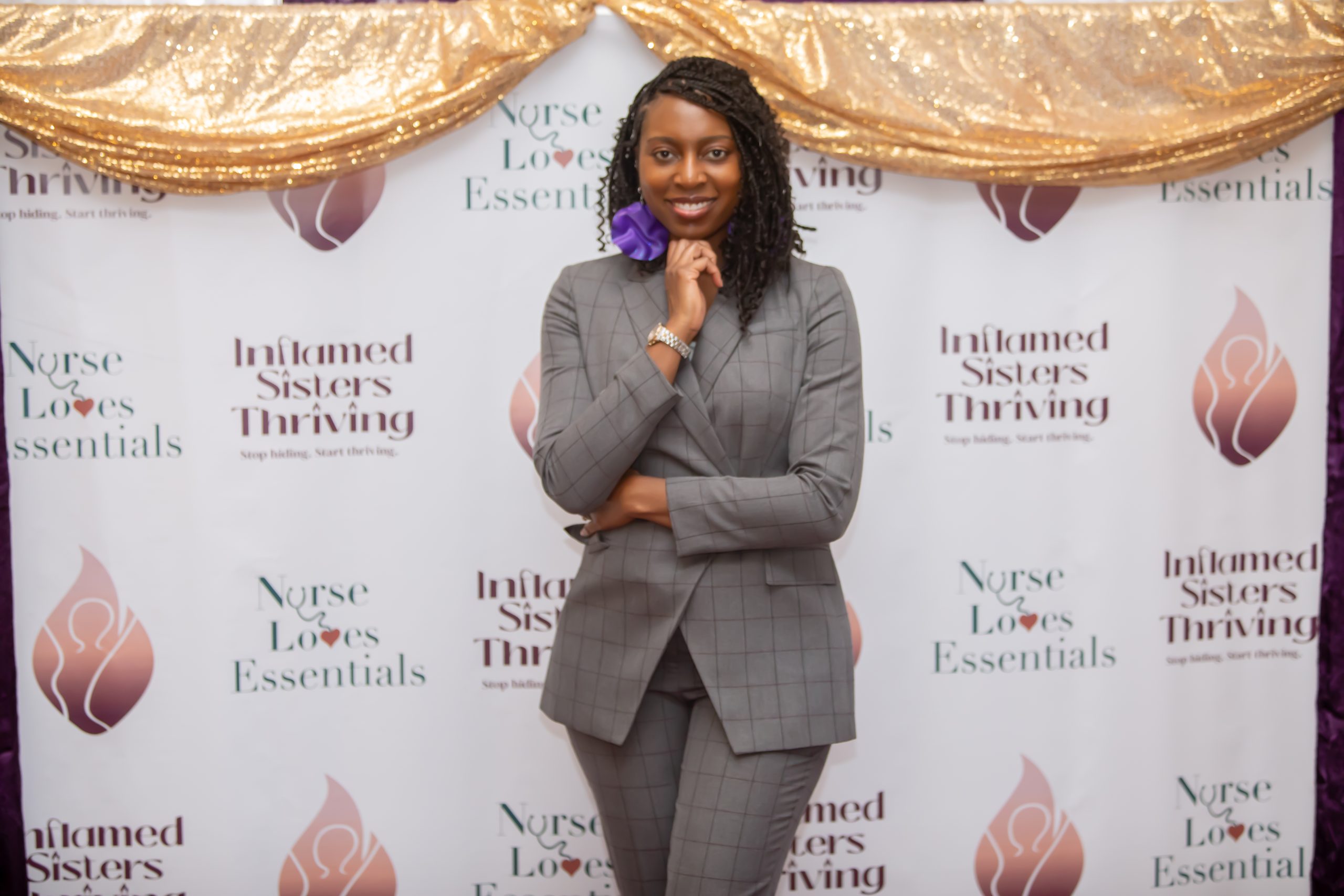 Catina Morrison, brown skinned professional woman with natural hair in grey suit with purple pinstripes standing by a banner with Nurse Loves Essentials LLC and Inflamed Sisters Thriving Inc.