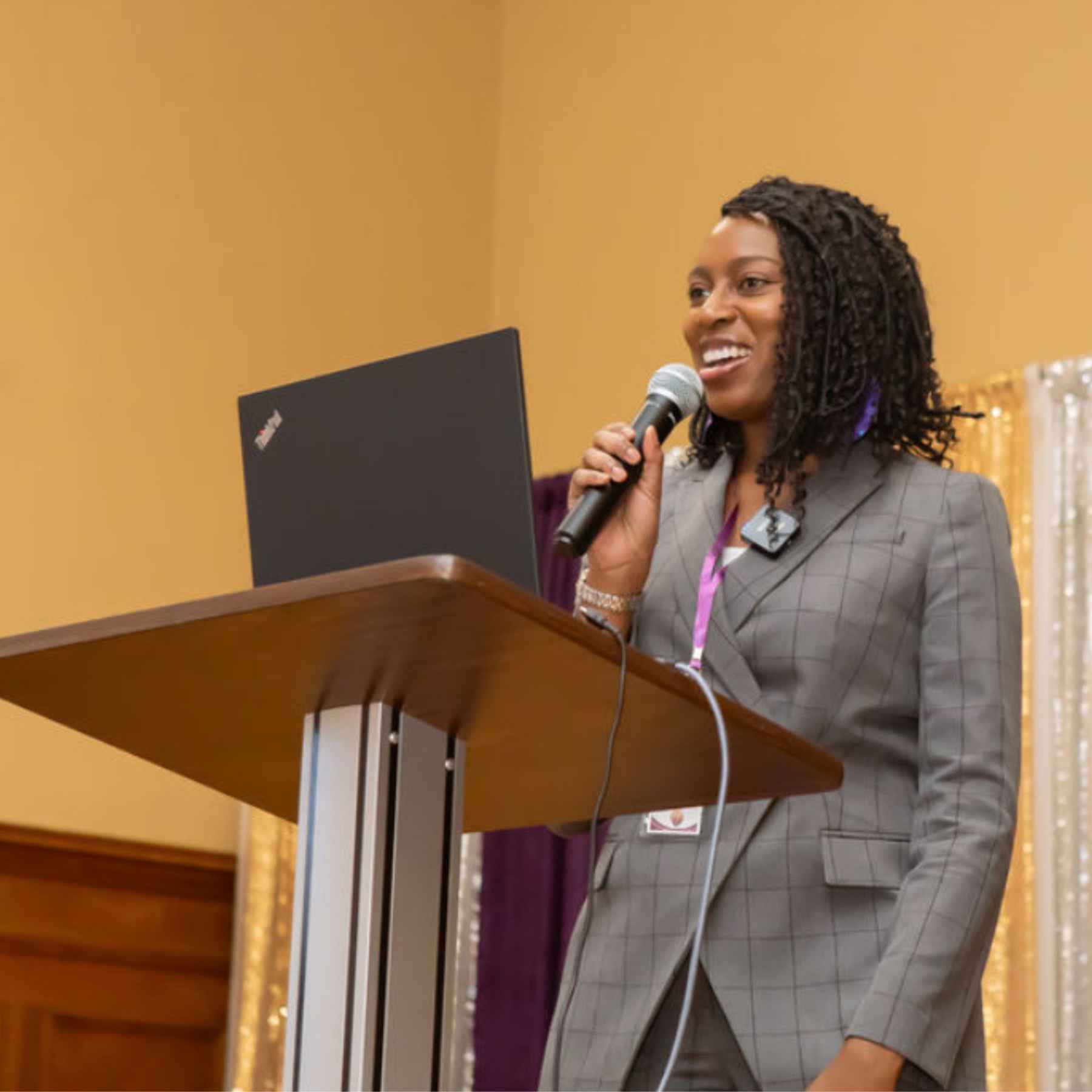 Catina Morrison, brown skinned professional woman with natural hair in grey suit with purple pinstripes speaking with a microphone in her hand while speaking at a podium.